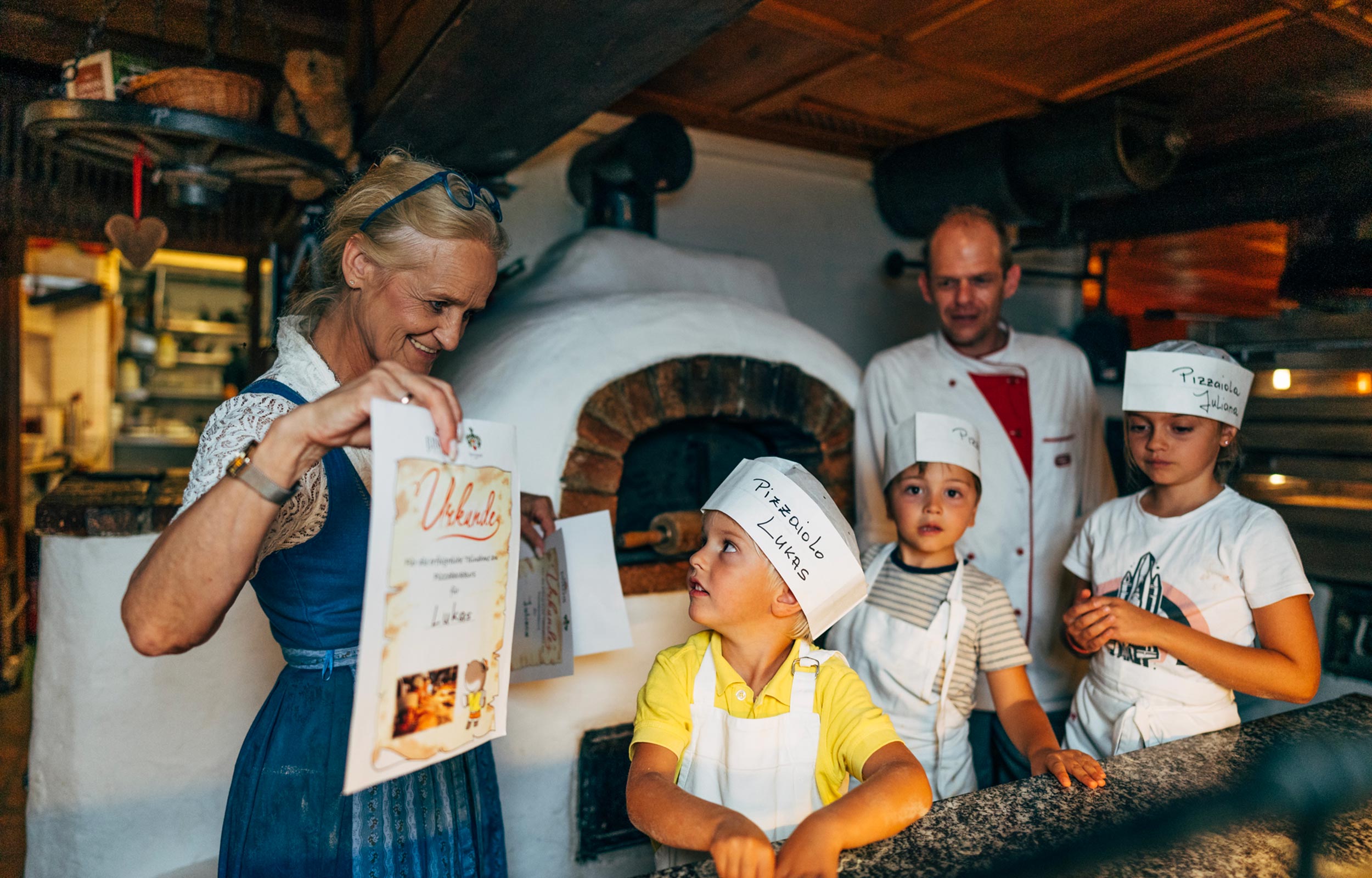 Three children wearing aprons and a medal receive a certificate for attending the pizza-making course at the Hotel Trattlerhof in Bad Kleinkirchheim